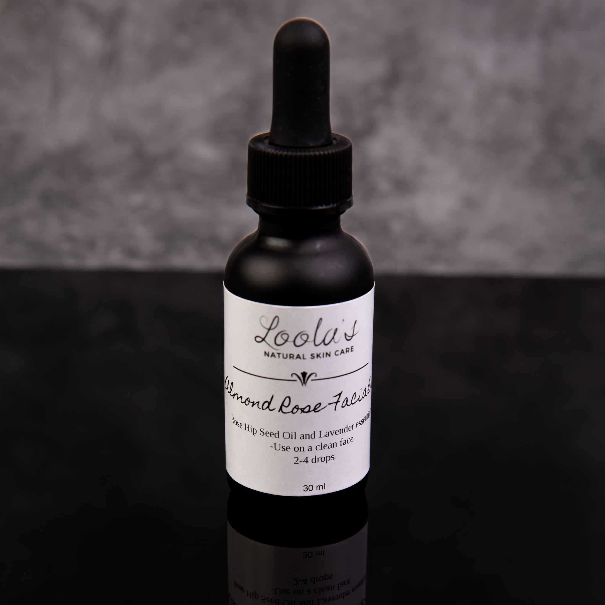  "Glass dropper bottle of Loola's Natural Skin Care Almond Rose Facial Oil, 30 ml, on reflective surface with ingredients listed as rose Hip Seed Oil and Lavender, against a grey stone background.